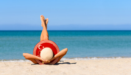 Wall Mural - A slender tanned girl on the beach in a straw hat in the colors of the flag of Turkey. The concept of a perfect vacation in a resort in the Turkey. Focus on the hat.