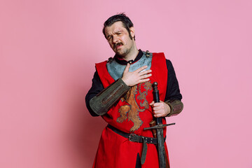 Wall Mural - One young man, medieval warrior or knight wearing wearing armor clothing posing isolated over pink background. Comparison of eras, history, emotions