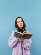 © bodnarphoto - Beautiful girl in a sweatshirt and headphones reads a book on a blue background with a serious face. Vertical
