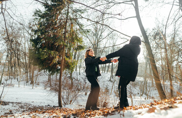 Two beautiful girlfriends in dark winter clothes have fun while walking in the snowy forest at sunset, holding hands and twirling with a smile on their faces.