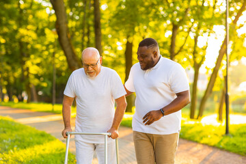 Caregiver is teaching old man to walk with walker. Professional nurse and patient walking outdoor at sunset. Assistance, rehabilitation and health care.