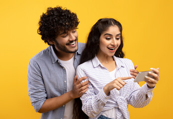 Wall Mural - Excited indian couple looking at smartphone screen, using new app or reading cool news, standing over yellow background