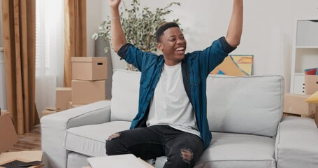 Wall Mural - A man sits on a couch by cardboard boxes left over from a move, happy with his new apartment, ordered furniture, joy, clapping hands