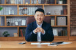 © Liubomir - Successful asian broker businessman looks at camera and smiles, man at work sitting at table in classic office