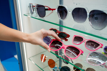 Close-up of woman choosing pink sunglasses on eyewear display stand in optical store. Woman taking frame from rack. UV protective eyewear concept.