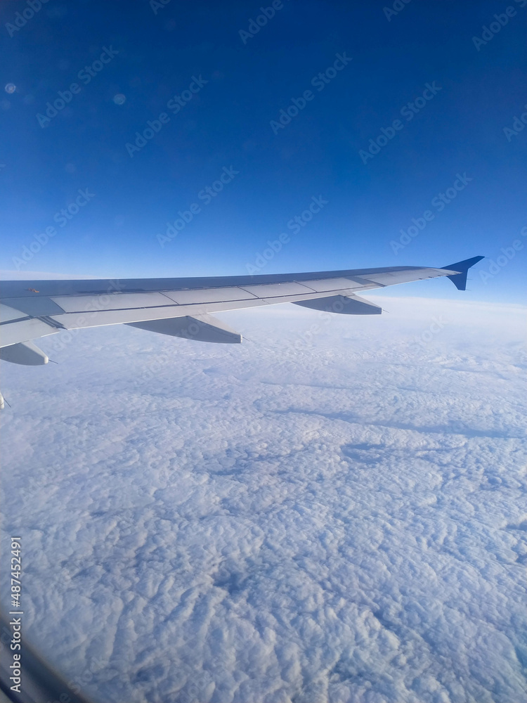 The view from the plane window on a cloudy sky in winter. Cabin window, snow, blue sky, plane wing, frozen plane window, flying. View on earth from a plane.
