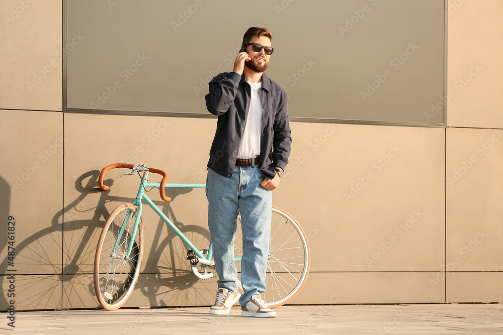 Stylish bearded man with bicycle talking by mobile phone in city