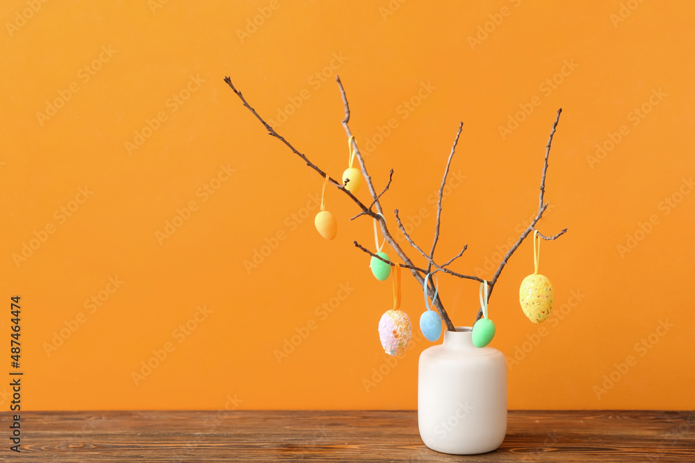 Vase with tree branches and different Easter eggs on wooden table against color background