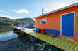 The exterior wall of an orange color wooden boathouse with a vibrant blue door, closed glass window, blue wooden benches, and long wharf with boats. 