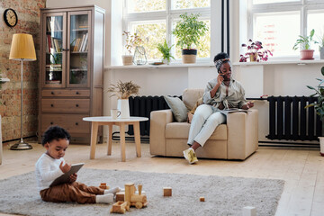 Wall Mural - Working single black mother answering call while her son sitting on floor and using tablet