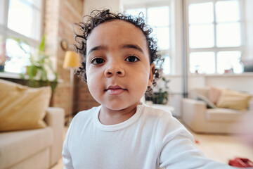 Canvas Print - Selfie of cute mixed race baby boy with dark curly hair wearing whute shirt sitting in living room