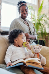 Canvas Print - Cute Afro-American toddler with curly hair sitting with book and toy bear in armchair and eating apple under control of mother