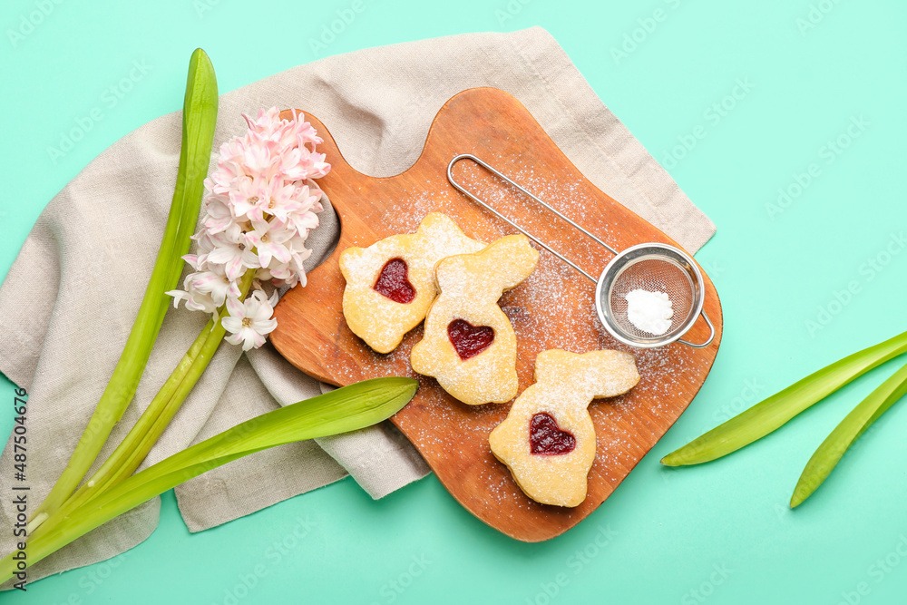 Board with delicious Easter cookies, sieve with flour and flower on turquoise background
