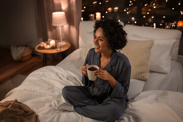 people, bedtime and rest concept - happy smiling woman in pajamas sitting in bed with hot chocolate mug at night