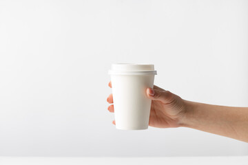 Human hand holding a white mockup closed paper coffee cup on a solid white background, morning in the coffeeshop, fast delivery craft fast food