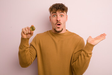 Wall Mural - young cool man looking surprised and shocked, with jaw dropped holding an object. bitcoin concept