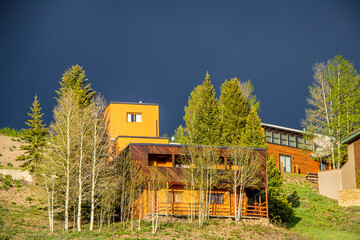 Modern wooden houses with view on slope surrounded by aspen trees under dark blue ominous sky in summer.