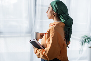 Wall Mural - side view of arabian student with notebook and tea cup looking away at home.