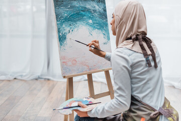 Wall Mural - young muslim woman in hijab painting on easel at home.