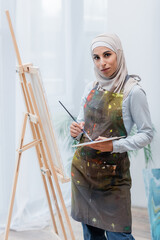 Wall Mural - arabian woman with palette and paintbrush looking at camera near easel.