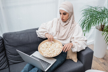 Wall Mural - arabian woman in hijab watching movie on laptop while sitting on sofa with bowl of popcorn.