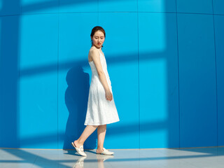 Beautiful Chinese brunette woman in white wedding dress dancing with her shadows on blue wall background. Sunny portrait of glamour young stylish lady. Emotions, beauty and lifestyle concept.