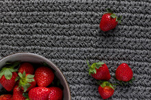 A Ceramic Bowl Full Of Fresh Strawberries Next To Another Four Isolated Strawberries Over A Gray Knitting Background.