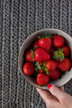 A Ceramic Bowl Full Of Fresh Strawberries Taken By A White Woman Hand Over A Gray Knitting Cloth Background.