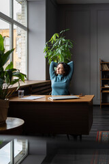 Wall Mural - Young relaxed businesswoman sitting at office desk, relaxing with hands over head, looking out window with dreamy face expression. Female employee taking break during busy workday, being happy at work