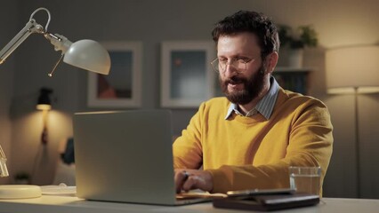 Happy man working at computer. Smiling laughing bearded man with glasses at home office sitting at desk and typing on laptop keyboard