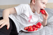 Child sitting on the couch and eating strawberry and spilling juice on t-shirt. The concept of cleaning stains on clothes. 