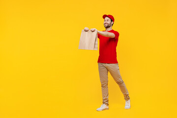 Wall Mural - Full body fun delivery guy employee man in red cap T-shirt uniform workwear work as dealer courier give brown clear blank craft paper takeaway bag mock up isolated on plain yellow background studio