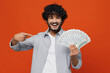 © ViDi Studio - Cheerful happy exultant young bearded Indian man 20s years old wear blue shirt holding pointing forefinger on fan of cash money in dollar banknotes isolated on plain orange background studio portrait