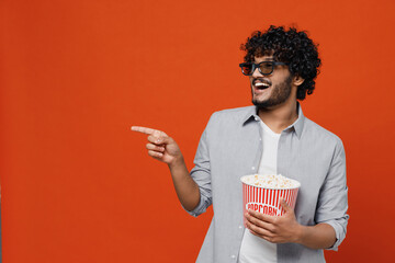 Excited exultant young bearded Indian man 20s wears blue shirt in 3d glasses watch movie film hold bucket of popcorn point forefinger away laughing isolated on plain orange background studio portrait.