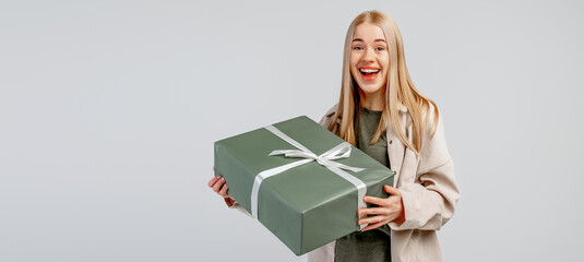 Portrait of Happy young blonde woman in trendy shirt holding big green gift box and looking at the camera over gray background. New Year Women's Day birthday holiday concept. Mock up copy space