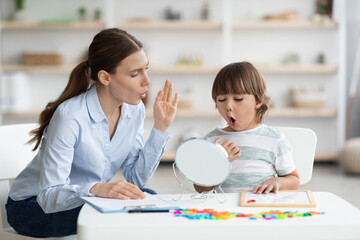 Wall Mural - Cute little boy pronouncing sound O looking at mirror, professional woman therapist teaching kid right pronounciation