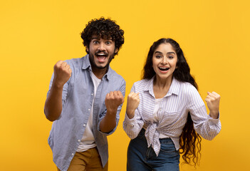 Portrait of overjoyed indian man and woman with open mouth cheering and staring at camera over yellow background