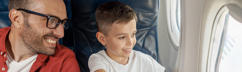 Wall Mural - Happy little boy looking out the window, showing something to his father while sitting near the window, traveling by plane together. Family, transportation, vacation concept