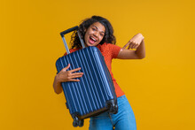 Studio Portrait Of Happy Smiling Tourist Girl Pointing At The Hand Luggage Bag She's Holding, Isolated Over Yellow Background