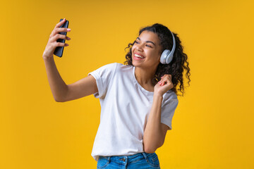 Wall Mural - Studio portrait of happy smiling beautful brunette girl wearing wireless headphones and taking selfie on her mobile phone, isolated over yellow background