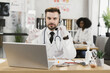 © sofiko14 - Confident medical specialist sitting at desk with syringe in hands and having online scientific discussion about vaccination with colleagues. Headset and laptop for conference call.