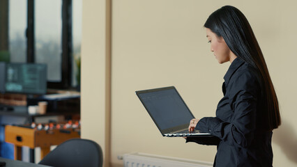 Asian software developer coming at desk and sitting down holding laptop with coding interface in cyber security agency. Programer getting ready to write code with colleague working in background.
