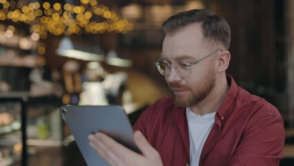 Skillful bearded programmer typing at the tablet while working at the cafe. Man using gadget with light smile. Technologies and people concept