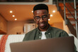 © Prins Productions - African American male sitting on sofa smiling while typing on laptop organising work for university