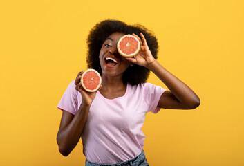 Summer mood. Overjoyed african american woman holding two grapefruit halves on yellow studio background