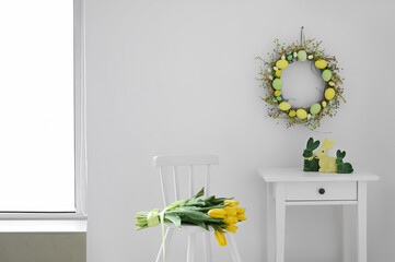 Bouquet of tulips and table with Easter rabbits near light wall with handmade wreath