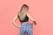 © Марина Демешко - A young caucasian slender blonde woman dressed in wide jeans stands with her back and demonstrates successful weight loss isolated on a light pink color background. Diet and wellness concept