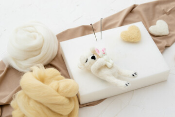 A small sleeping woolen white teddy bear made by dry felting. Felt toy for a newborn photo session. handmade product. Workplace for dry felting with a foam sponge, needles, fabric and skeins of wool 