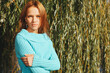 © Sergios - Beautiful redhead young woman posing on a bright sunny day against the background of willow branches in the park, dressed in a blue sweater. People and lifestyle concept