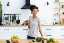 Healthy Diet Eating. African American Young Female Preparing Salad For Dinner Or Breakfast In Home Kitchen. Woman Cooking Healthy Food With Glass Of Clean Water, Looking Away, Smiling Friendly